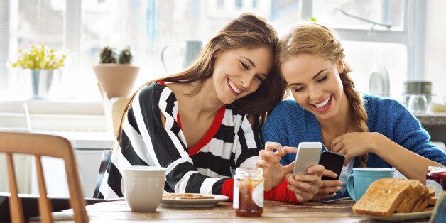 Two cheerful friends sitting at the kitchen table and using smart phones.