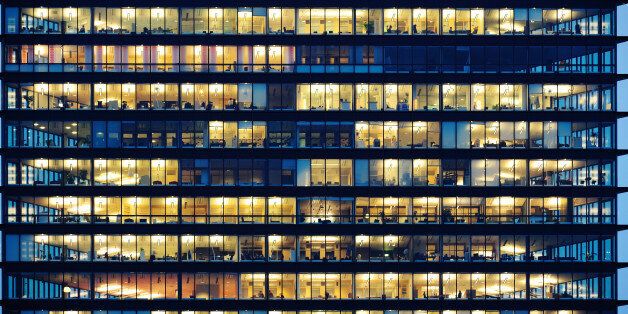Lots of people working late. Employees seen as silhouettes against their brightly lit offices with large windows. Building framed by the 'blue hour' evening sky