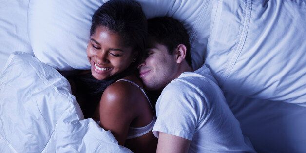 Photo of an attractive young mixed race couple, lying in bed as husband playfully invades his wife's side of the bed.