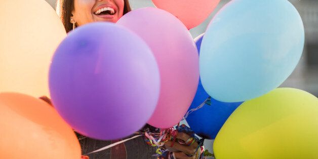 Cheerful woman peeking behind large group of multi-colored balloons and looking at the camera.