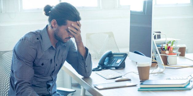 Stressed business man at the office. He is casually dressed and looking distraught. He looks very uncomfortable and could also have a headache. He is has his head in his hand and looks very upset. Hi is sitting at a desk with a computer and phone. Copy space.