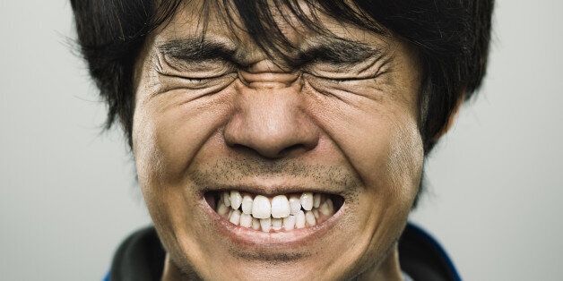 Studio portrait of a japanese young man with closed eyes and clenched teeth. The man has around 30 years and has long black hair and casual clothes. Vertical color image from a medium format digital camera. Sharp focus on eyes.