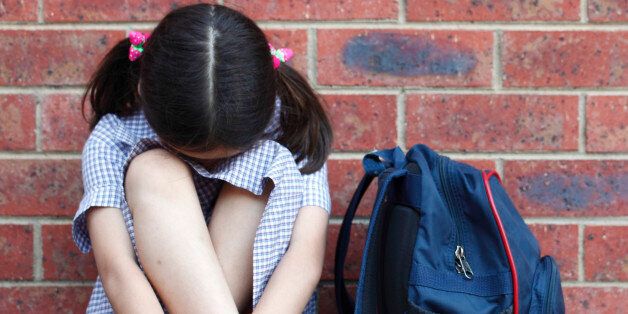 School girl. Tough times. Concept for bullying or challenges with growing up. A young girl wearing her school uniform sits with her head down. She is against a brick wall and her school bag sits alongside her.