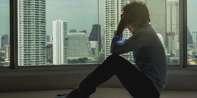 depressed man sitting head in hands on the interior Skyscraper with low light environment beside the windows over the cityscape background, dramatic concept
