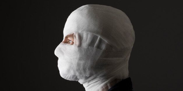 Injured young man after surgery with bandage all over his face with one eye opened in dark. Image related with treatment of the wounds, plastic surgery, medical industry