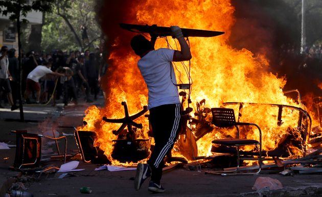 An anti-government protester adds an object to burning chairs and benches amid a march by students and union members in Santiago, Chile, Monday, Oct. 21, 2019. Protesters defied an emergency decree and confronted police in Chile’s capital on Monday, continuing disturbances that have left at least 11 dead and led the president to say the country is “at war.” (AP Photo/Miguel Arenas)