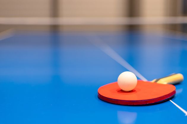 A closeup of a red table tennis paddle with a wood handle and ping pong ball on a blue table.