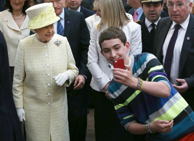 A local youth takes a selfie photograph in front of Queen Elizabeth II during a visit to St George's indoor market on June 24, 2014 in Belfast, Northern Ireland.  AFP PHOTO/POOL/PETER MACDIARMID        (Photo credit should read PETER MACDIARMID/AFP via Getty Images)