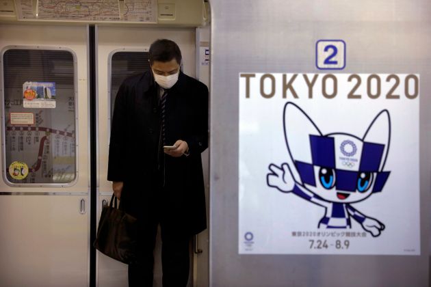 A poster promoting the Tokyo 2020 Olympics is posted next a train door as a commuter wearing a mask looks at his phone in a train, Friday, Jan. 31, 2020, in Tokyo. Tokyo Olympic organizers are trying to shoot down rumors that this summer's games might be cancelled or postponed because of the spread of a new virus. (AP Photo/Jae C. Hong)