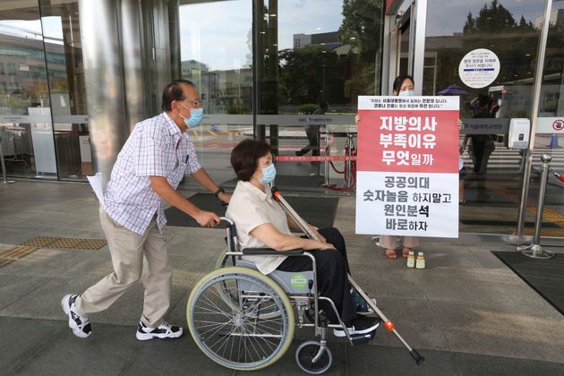 8월 26일 수요일 한 의사가 서울대학교병원에서 정부의 의료정책을 비판하는 팻말을 들고 있다. 코로나바이러스 환자가 하루 연속 3배 이상 증가한 것으로 집계됨에 따라 한국 보건 관계자들은 수천 명의 파업 중인 의사들에게 업무에 복귀할 것을 요구했다. 표지판에는 다음과 같이 쓰여 있다: '이 지역의 의사들이 부족한 것은 무엇인가?